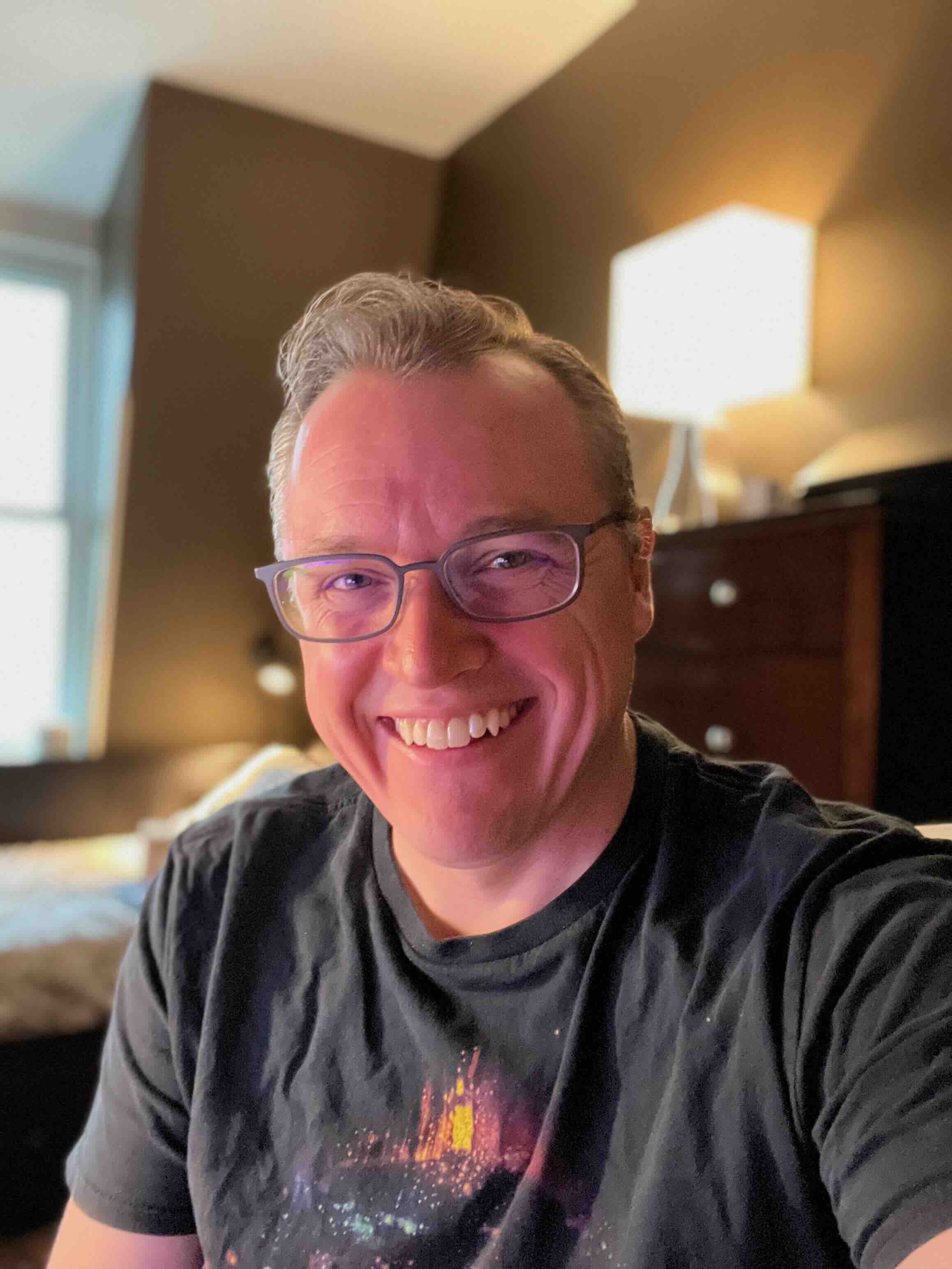 Smiling person with glasses wearing a black t-shirt, standing in a room with soft lighting and modern decor.